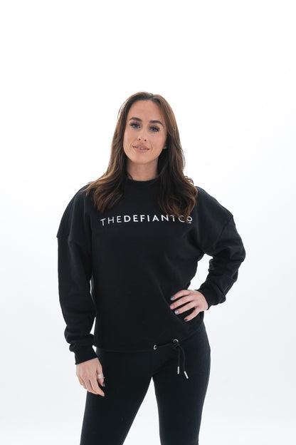 A photo showing The Defiant Co women's oversized crewneck. The crewneck has a roundneck and draw string around the bottom. The crews have the famous The Defiant Co logo across the front of the chest and are plain on the back. The colour is black.