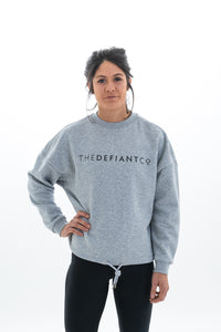 A photo showing The Defiant Co women's oversized crewneck. The crewneck has a roundneck and draw string around the bottom. The crews have the famous The Defiant Co logo across the front of the chest and are plain on the back. The colour is heather grey.
