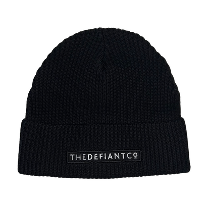 A photo showing a beanie hat. The hat has a ribbed finish with a roll cuff.  The cuff has The Defiant Co logo embroidered on the centre of the front in white on a black background. The beanie hat is black.