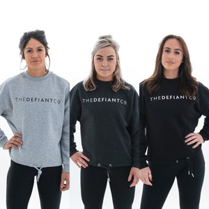 A photo collage showing the three variations of The Defiant Co women's oversized crewneck. They crewnecks have a roundneck and draw string around the bottom.  The crews have the famous The Defiant Co logo across the front of the chest and are plain on the back.  The colours available are heather grey, charcoal and black.